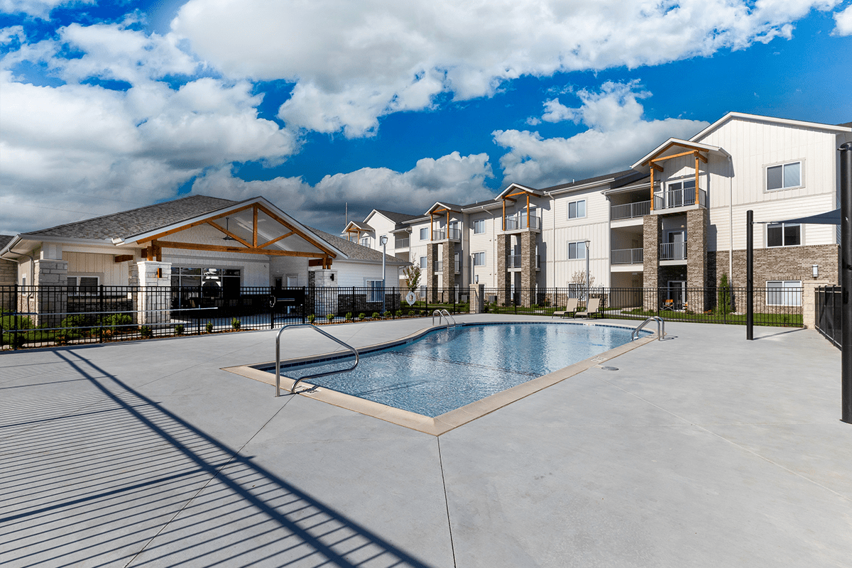 A swimming pool with lounge chairs and a fence surrounding the area with apartments in the background.